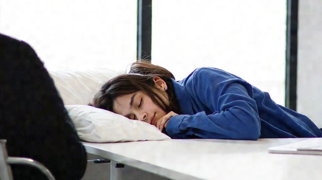 A young woman wearing a blue shirt appears to be sleeping on a desk during the daytime, with her head resting on a pillow. The setting resembles a workplace or classroom environment, highlighting the possible challenges of managing narcolepsy, a sleep disorder characterized by excessive daytime sleepiness and sudden sleep attacks.