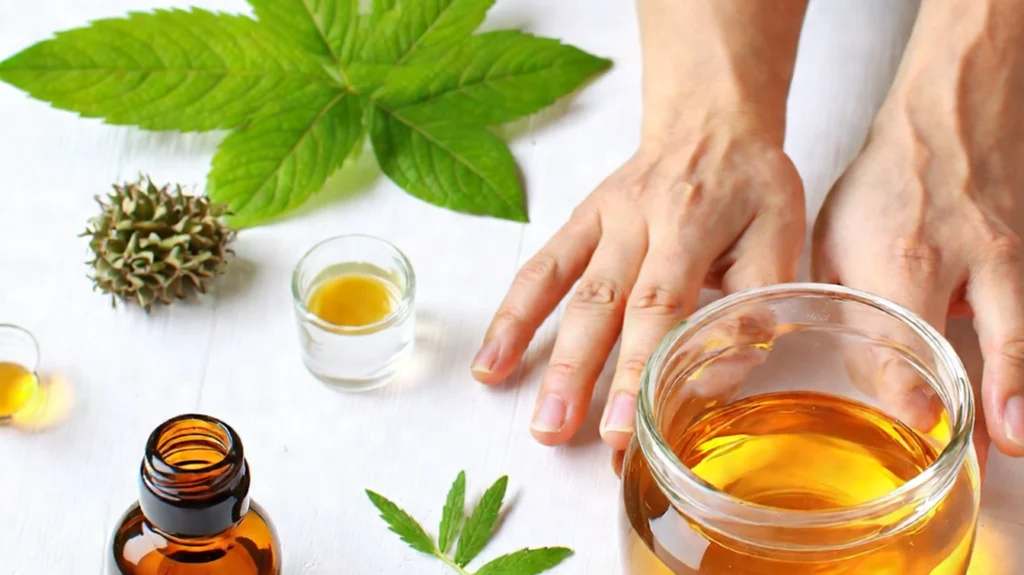 A natural remedy setup for headaches, featuring a jar of golden oil, small glass containers of oil, a spiked seed pod, and fresh green leaves on a white wooden background. The scene also includes a pair of hands gently placed near the jar, suggesting preparation or application of the remedy.